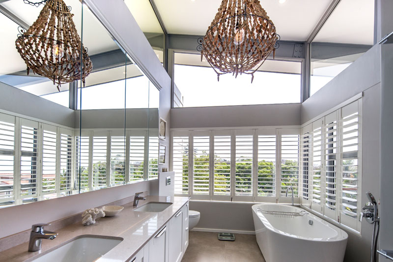 In this modern bathroom a free standing bathtub is situated by windows that are covered by white shutters. Clerestory windows add extra light to this airy washroom. 