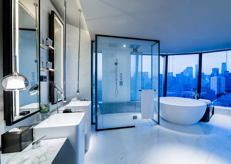 In this modern hotel bathroom, a white standalone tub sits in front of the glass enclosed large shower, and faces the city of Beijing. The bathroom decor is minimal, allowing the city view to be the main decorative element.