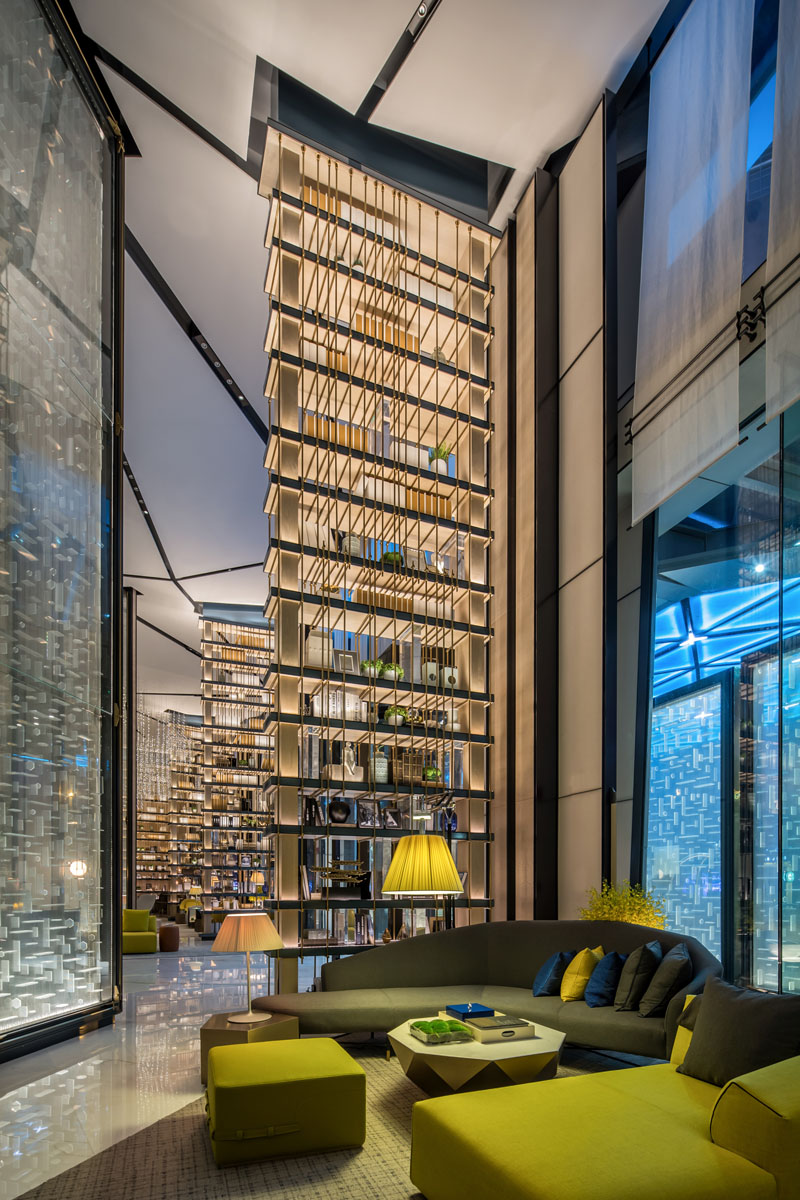 In this modern hotel, huge vertical partitions and custom-made sofas line the long narrow hallway of the modern lobby, separating spaces from one another. The shelves of the partitions are lit, and are luxuriously decorated with various ornaments.