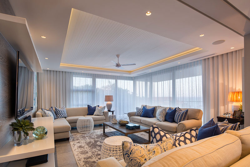 This modern living room has floor-to-ceiling windows in the corner along with a recessed ceiling with hidden lighting makes this already light room brighter. Decorated in blues, yellows, and grey, the living room brings tones of the beach from outside to inside. 