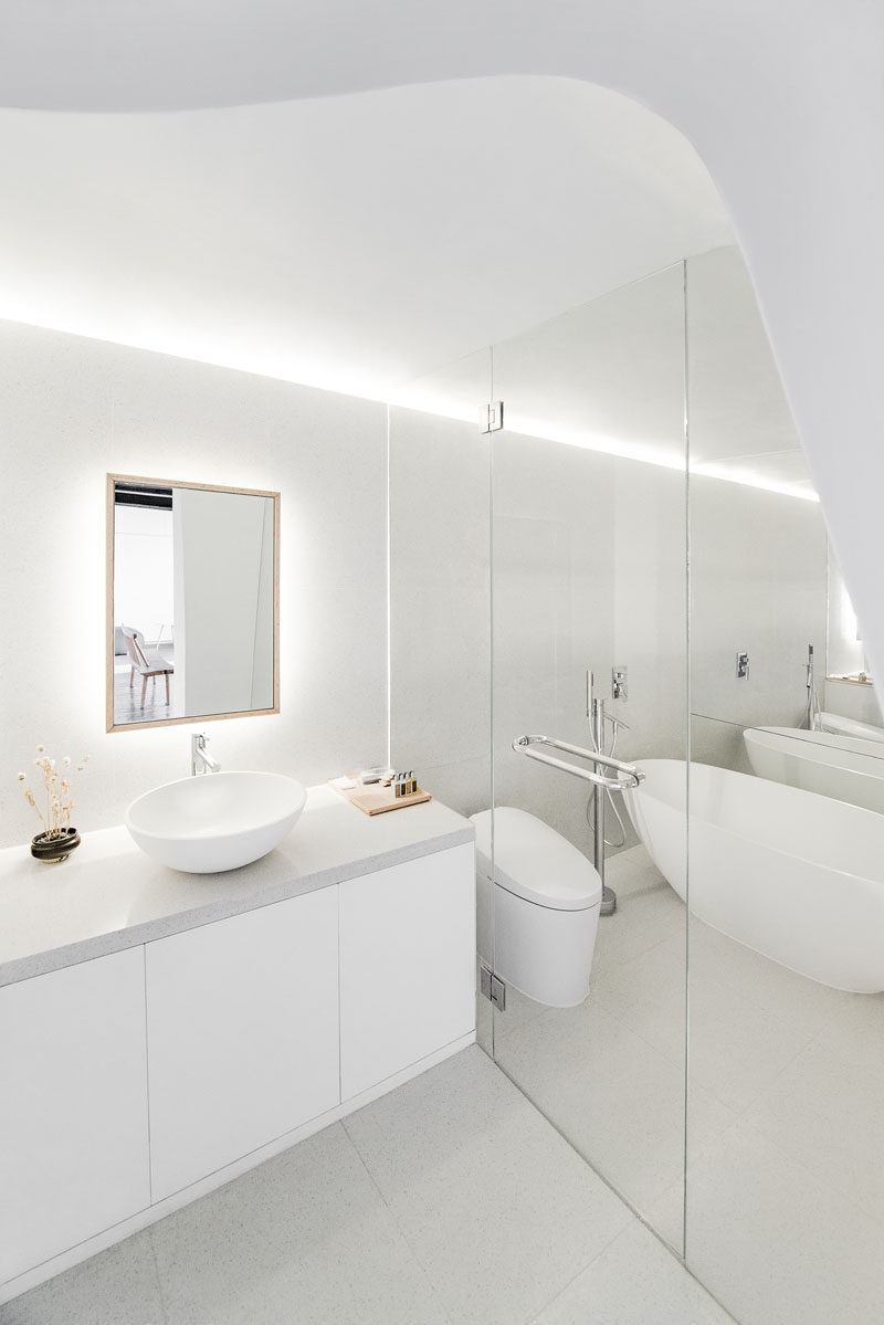 In this modern all white bathroom, there's a curved ceiling element and hidden lighting in the ceiling. A back lit mirror makes this bright room even lighter.