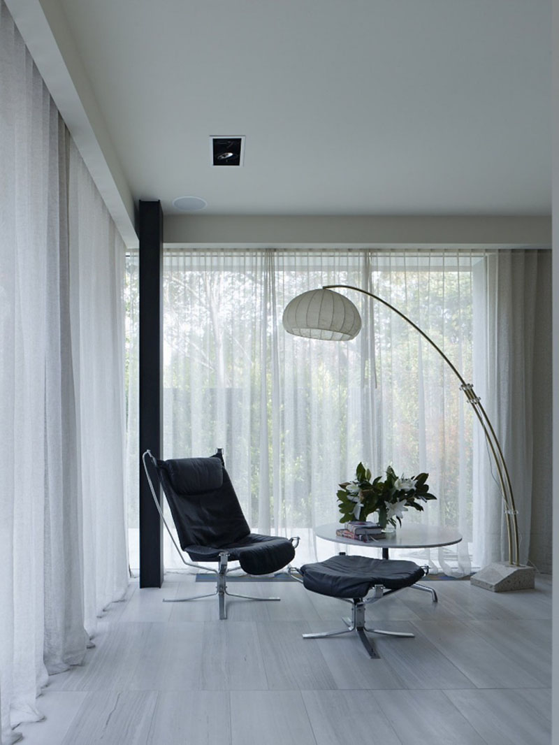 Stepping inside this modern home, a large floor lamp with a white shade provides light above a black upholstered leather chair and a small table. 