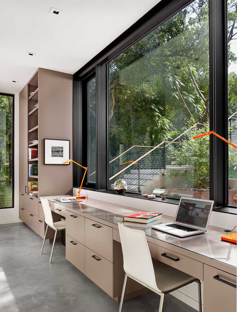 The light colored cabinets, shelves, and desk for two, is built-in to the wall of this modern office. Facing a large black framed window, this work space makes it hard not to day dream.