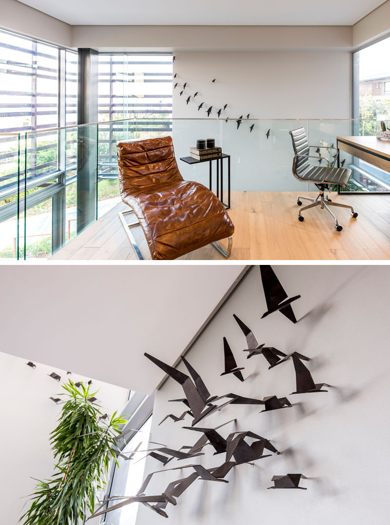 Surrounded by glass railings, this modern home office has a light wood desk that faces a window, and a brown leather chaise lounge for reading. A 3D steel art installation of flying birds, designed by the home owner, decorates the far wall.