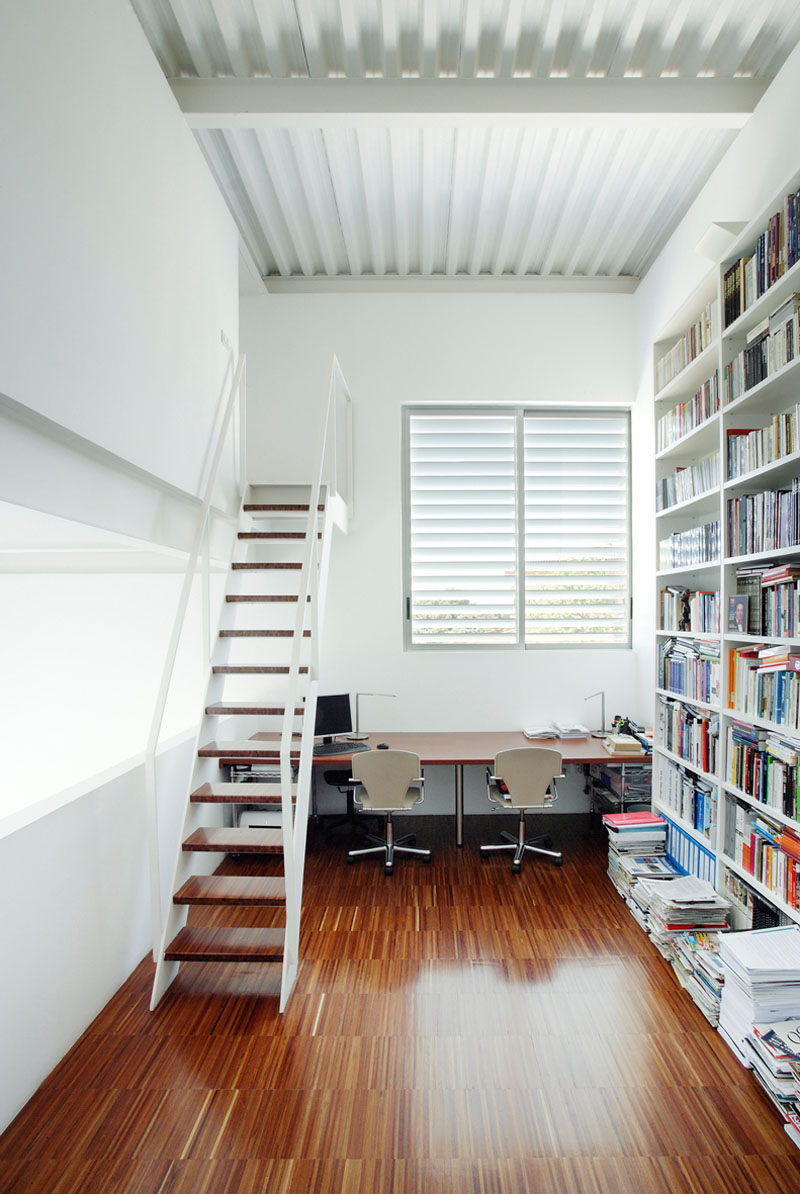 Almost entirely white, this office for two has a near floor-to-ceiling bookcase, and a set of stairs that lead to another room. The wood desk below the white framed window, matches the floor and stairs.