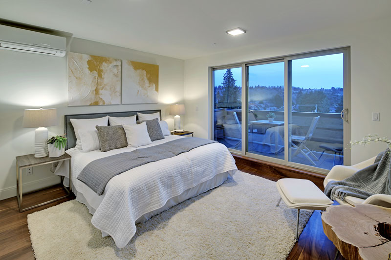 In this modern bedroom the bed sits beside a set of sliding glass doors that lead to a small balcony. A white plush area rug on the walnut floor gives the room a cozy feel.