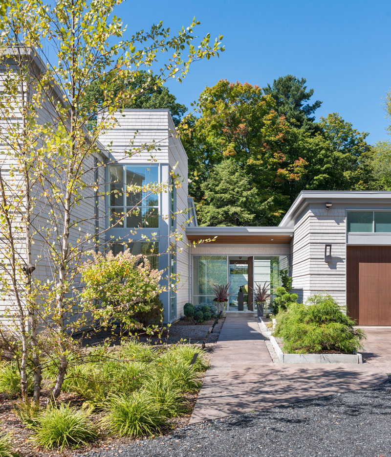 A landscaped path leads to the front entrance of this modern house, with the garage on the right and the house on the left. The home has been designed with a flat roof and floor to ceiling glass windows, allowing sunlight to fill the interior.