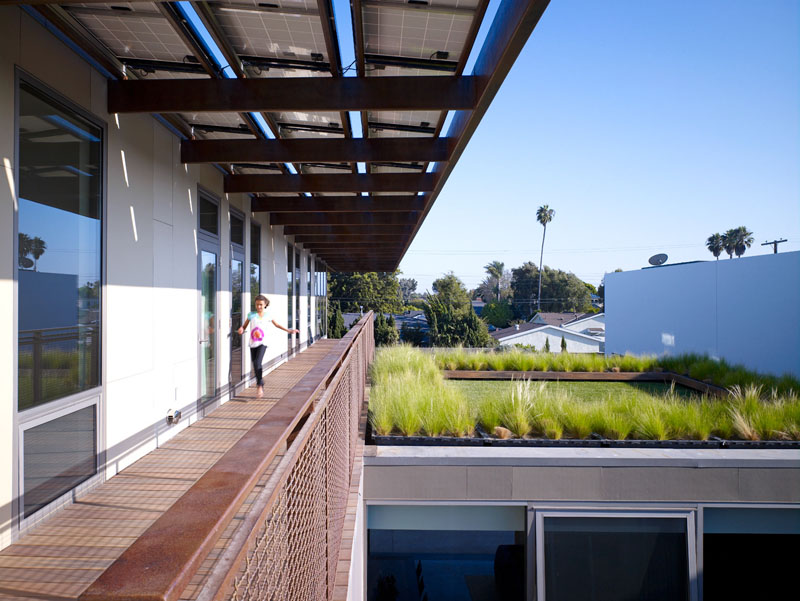 This modern long exterior walkway appears at the top of this house, leading to the bedrooms. A green roof has been added above the living room to provide environmental benefits.