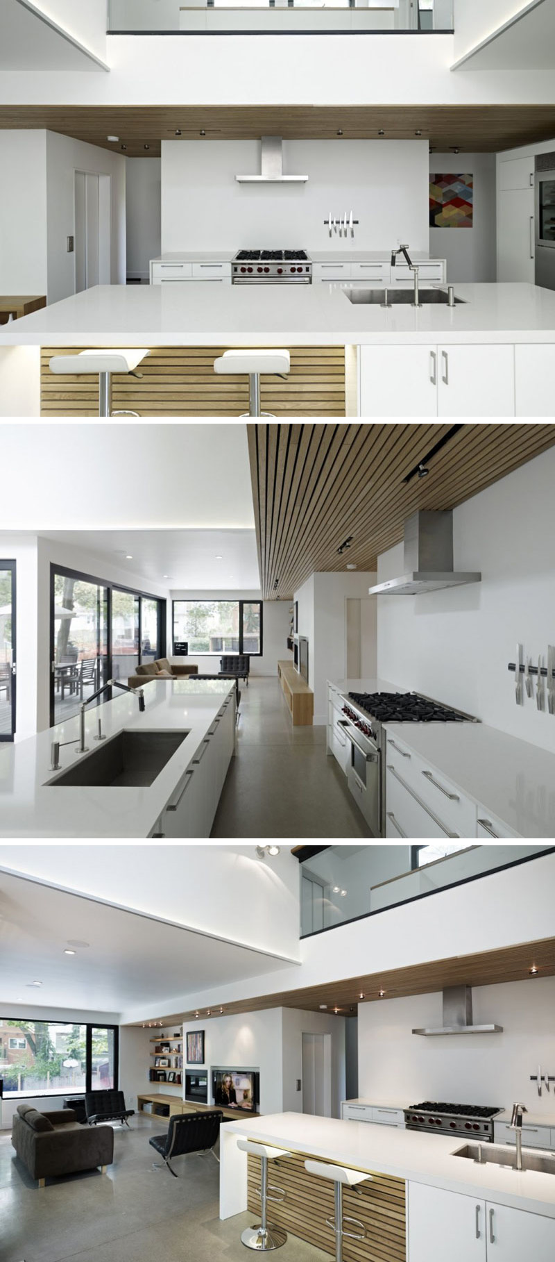 In this modern kitchen a long white island with wood accents has a built-in sink and plenty of storage. Above the stove, a slat wood accent feature in the ceiling with lighting continues into the living room.