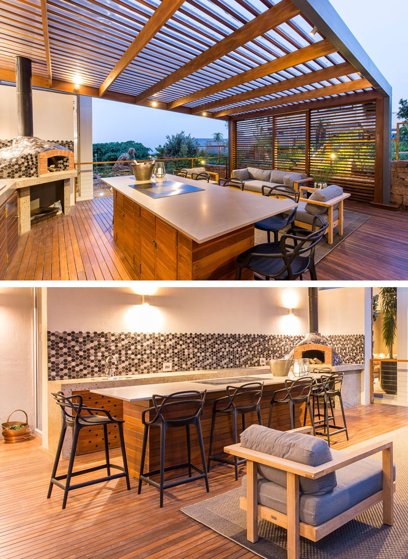 This modern outdoor entertaining space has a wood pergola, an outdoor kitchen and a lounge area. A wood island with a grey countertop provides space for cooking and eating, while a prep area with a patterned tile backsplash can be used to make pizzas in the wood fired oven.
