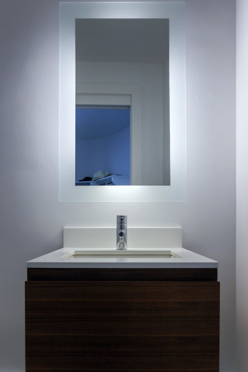 In this modern bathroom, a white sink with wood cupboards sits below a back-lit rectangular mirror with opaque borders.