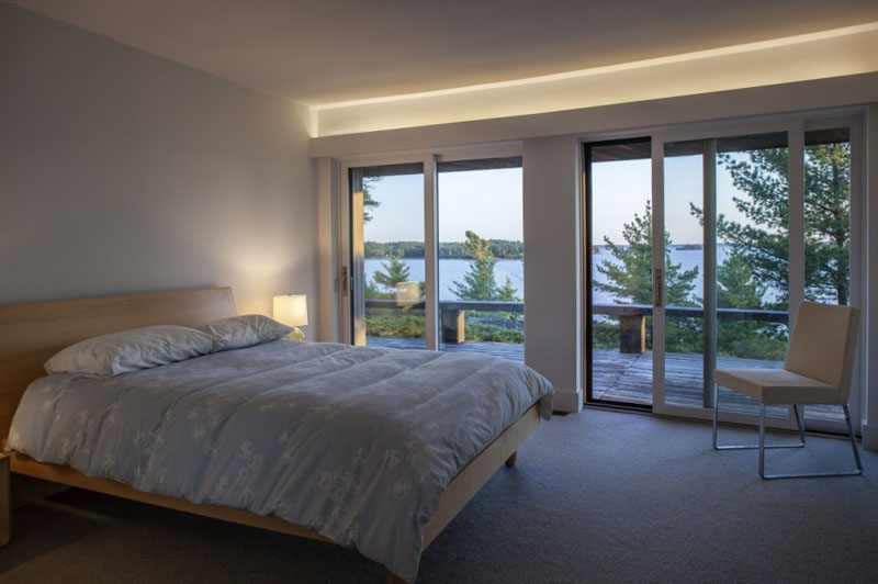 In this modern bedroom, a wood framed bed sits beside a set of glass sliding doors that lead to a balcony. Hidden lighting is used in this room above the balcony doors. 