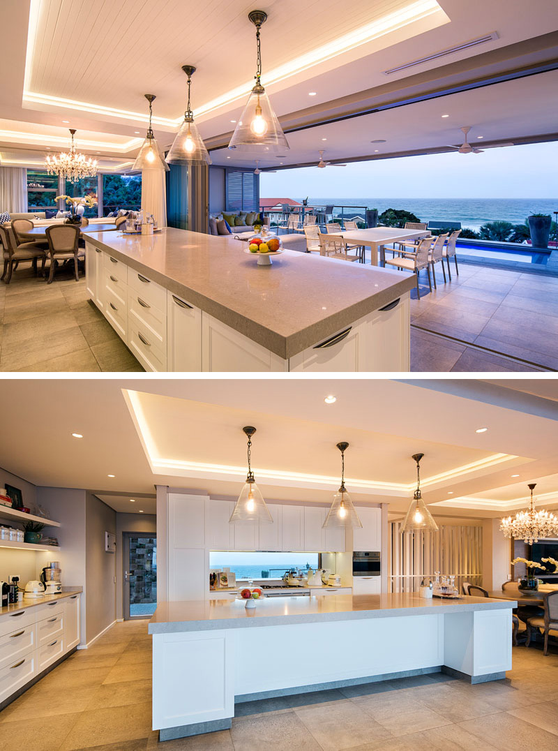 In this modern kitchen, a long white and grey island sits below three glass pendant lights that are hung from a recessed ceiling with hidden lighting. The kitchen faces the pool and views the expansive ocean. 