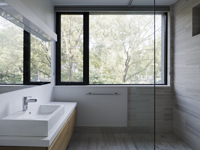 In this modern bathroom, a glass surround shower is placed beside a large black framed window providing a view of outside. A wood vanity with white counter and sink adds extra storage to the bathroom.