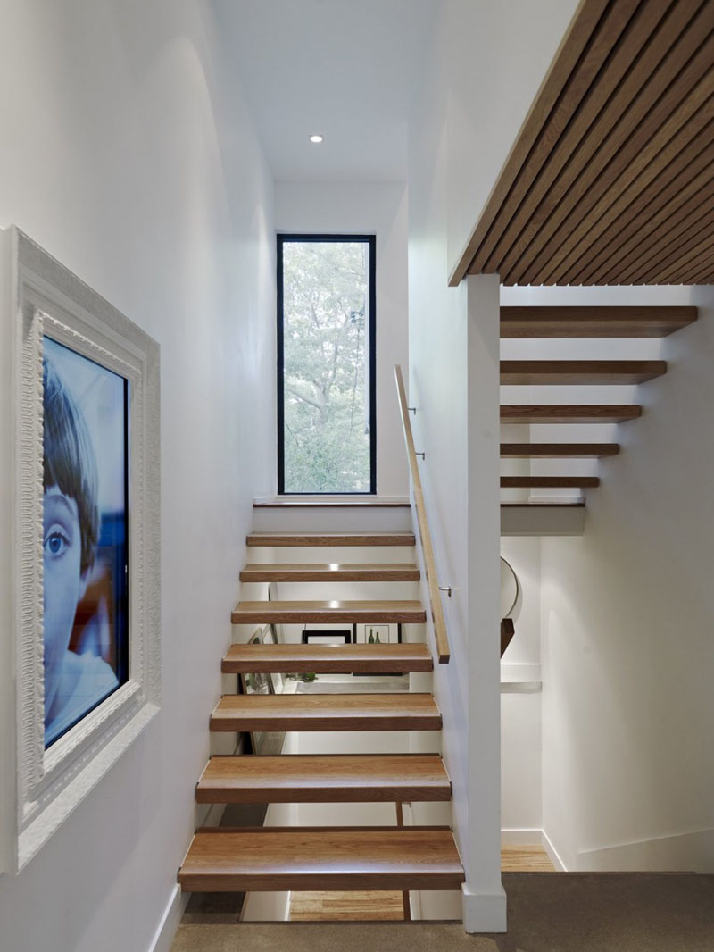 This modern set of wood stairs with a wood hand railing lead to the upper floor of the home. A large white framed digital picture decorates the wall, while a vertical window provides glimpses of the trees outside.