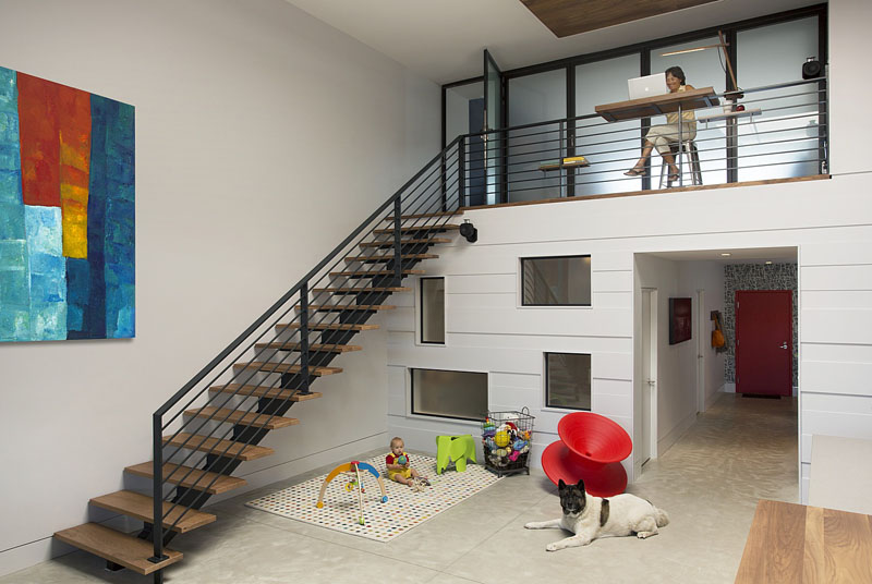 In this modern loft, walnut wood stairs with a black hand railing lead to the lofted part of the home. Underneath the stairs, a small area has been set up as a play area, and you can see the windows that look into the nursery.