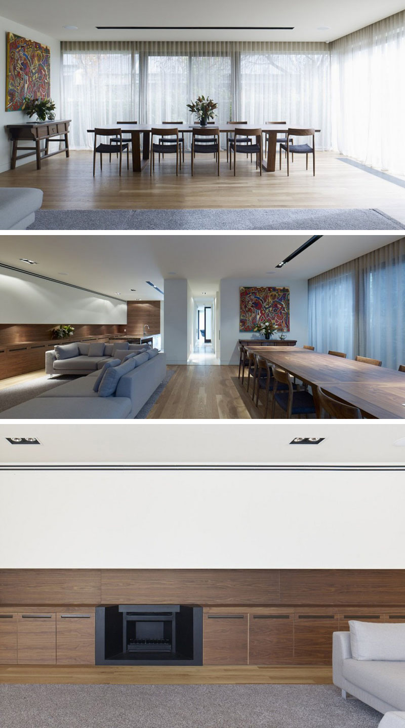 In this modern dining area, a long wood table is featured surrounded by floor-to-ceiling windows that are covered by sheer white curtains. On the opposite side of a large grey upholstered sofa is the living room which has dark wood cabinets. 