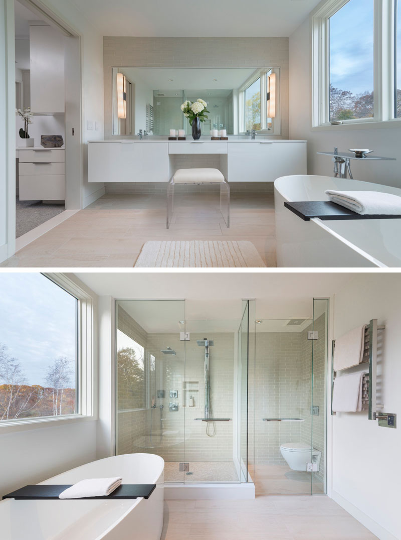 In this modern master bathroom, a large rectangular mirror above the white counter has long opaque lights attached to either side. The glass surround shower is placed beside the toilet which is also surrounded by glass. 