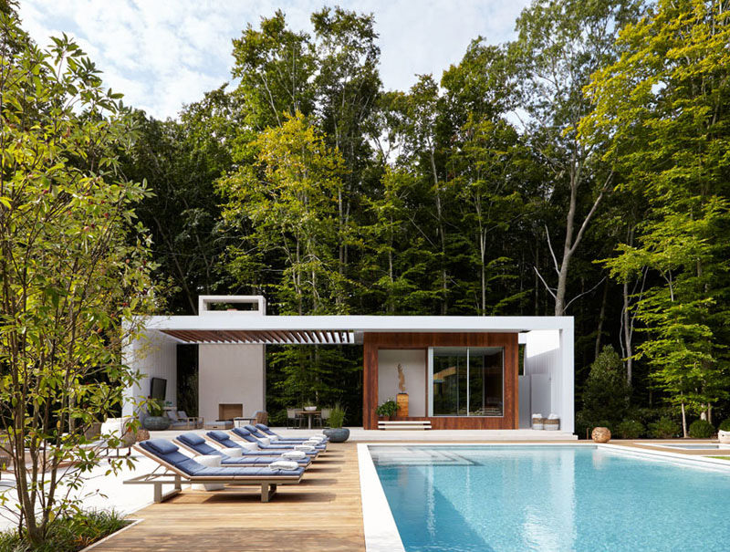 This white and wood pool house features a covered outdoor lounge area with a fire place. Blue upholstered sun chairs line the length of the pool.