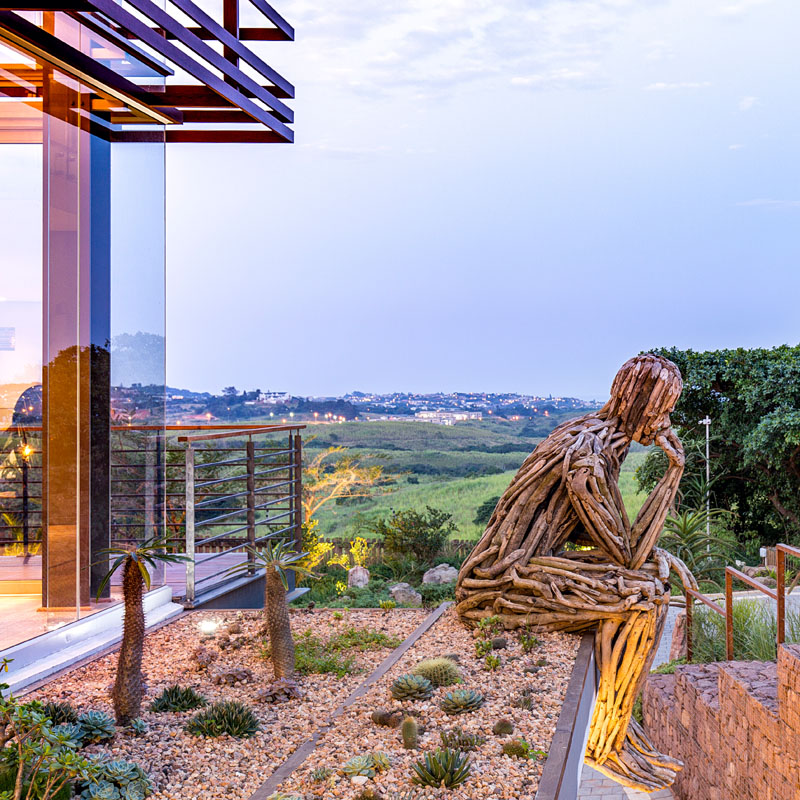 Another large wood art installation, the thinking man, sits in front of a mini rock garden and looks out over the side of this modern house.