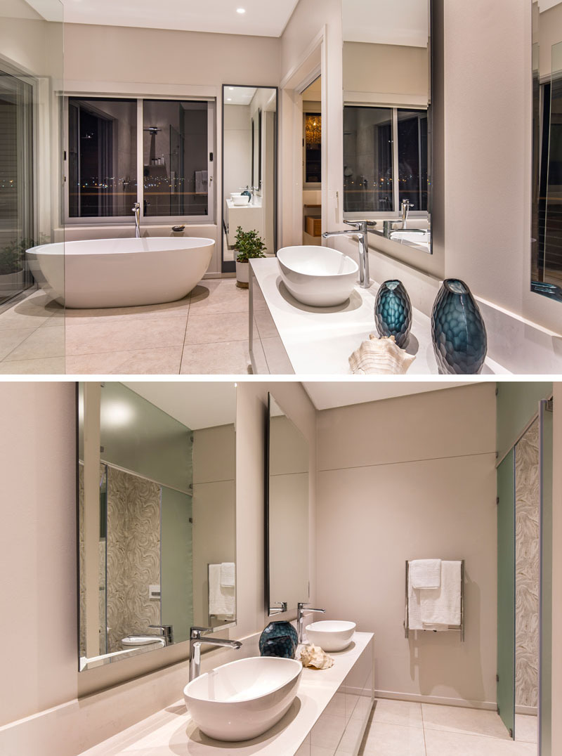 In this modern master bathroom, a white standalone tub is placed beside a large window providing a view of the city. In front of the glass shower, his and her sinks sit on top of the white counter.