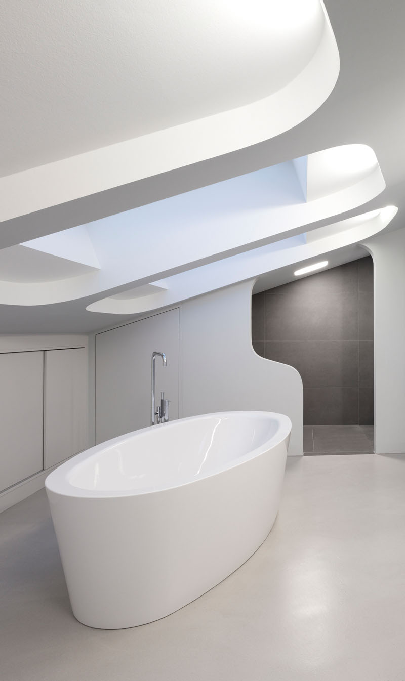 This modern bathroom has a white standalone bathtub that sits below a large, recessed skylight. Rectangular grey tiles are used in the portion of the bathroom where the toilet is. 