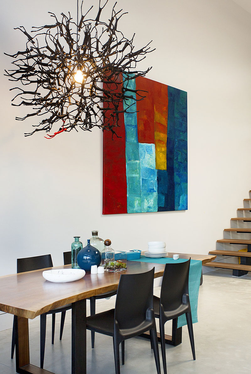 In this modern dining area, a wood dining table sits below a sculptural pendant light made from human-like figurines. A wall mounted painting with reds, blues, yellows, and whites ties in the various colors used throughout this modern loft interior.