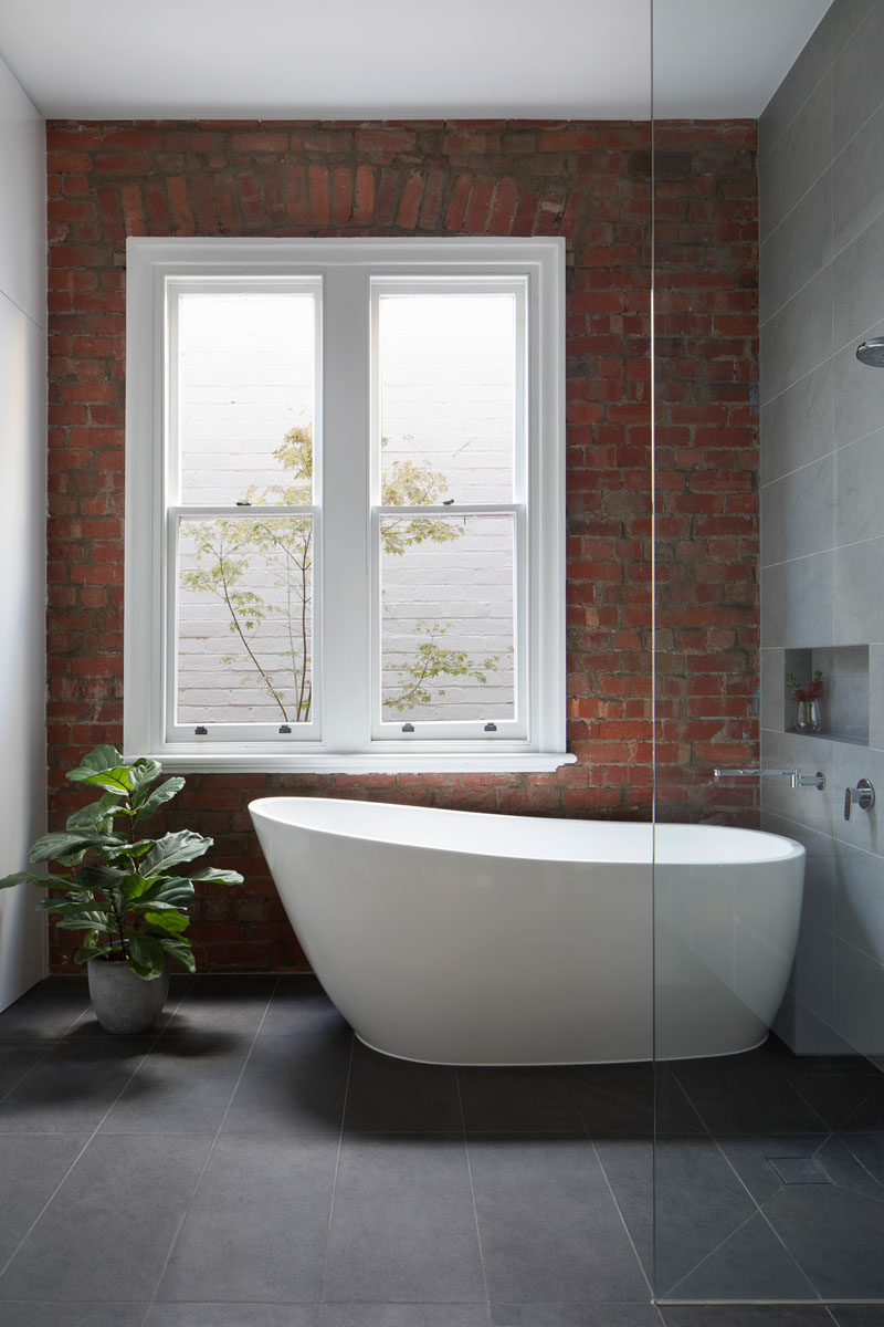 In this updated bathroom with large dark grey tiles and a white freestanding bathtub, the heritage parts of the house were retained, like the brick wall that's been left on display instead of covered up. 
