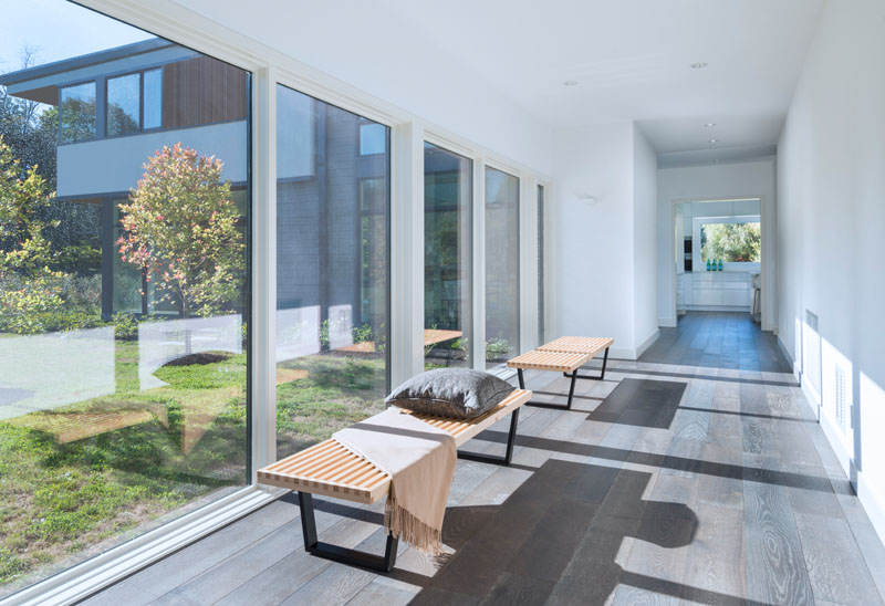 Going inside this modern house, two wood benches are placed in front of large windows that line a long white hallway with dark wood flooring.