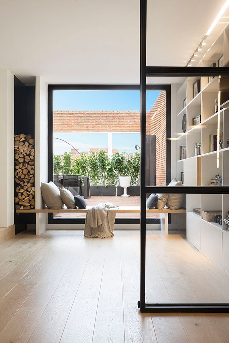 A built-in wood window seat sits between the shelving unit and the firewood storage in this modern penthouse, while the large window provides plenty of natural light and views of the outdoor space.