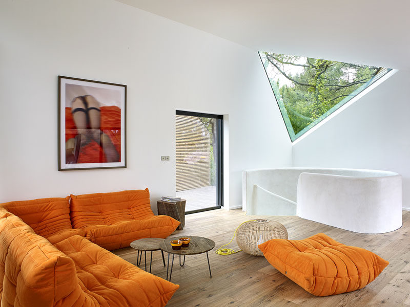 In this modern living room, the bright orange couch creates a comfortable spot to sit, while a triangular window provides a unique view of the trees.