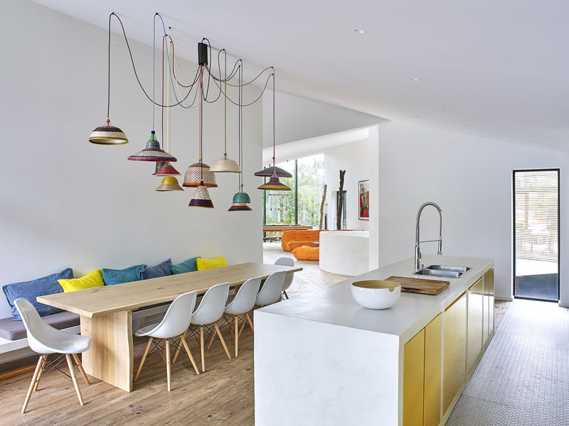 In this modern holiday house, the dining area and kitchen share the same room. The large dining table is anchored in the room by a collection of colorful pendant lights hanging from the ceiling. In the kitchen the cabinets have a gold metallic front, adding to the touch of color in the white space.