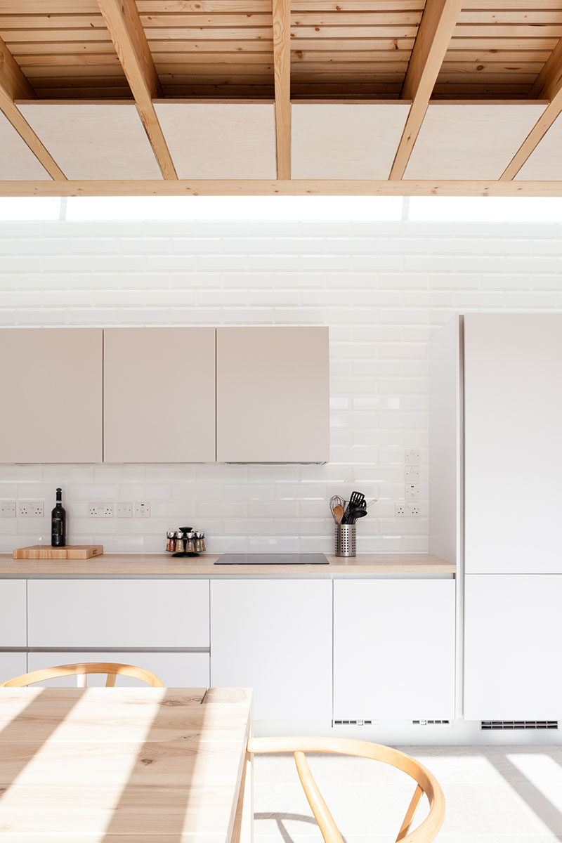 Inside this modern house extension is a kitchen with white and tan cabinets that's positioned along the wall, while white tiles help to reflect the light from the skylight above the kitchen.