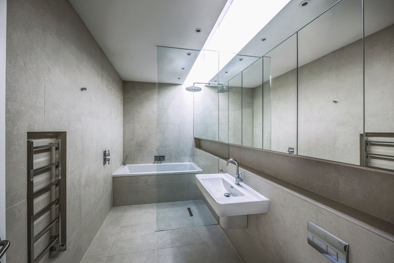 A skylight has been installed in this modern bathroom, creating a bright strip of natural light that brightens up the neutral color palette. A wall of mirrors runs the length of the room and a glass partition separates the wet areas from the rest of the bathroom.