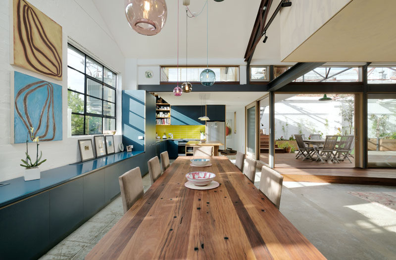 In this modern converted warehouse, custom blue cabinetry runs along the wall and into the kitchen, while a large wood dining table is anchored in the space by colorful glass pendant lights.