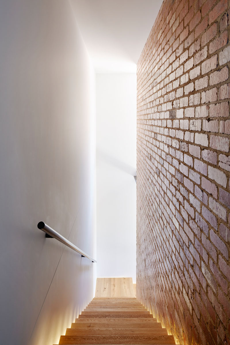 Wood stairs, sandwiched between a white wall and a brick wall, are lit up with hidden lighting on either side.