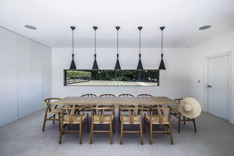 In this modern and formal dining room, five black pendant lights hang above wood table and chairs, while a large horizontal window perfectly frames the garden.