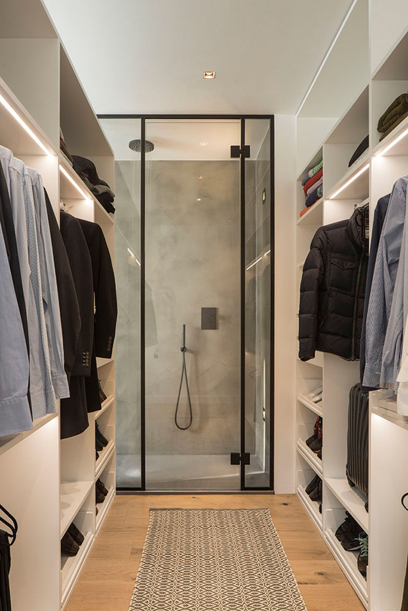 This modern walk-in closet has a black framed, glass shower at one end, while some of the shelves in the closet have strips of lighting to add additional light to the space.