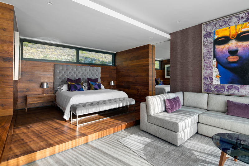 In this modern master bedroom, the bed sits slightly raised on a wood platform, which a grey couch provides a nice spot to relax.