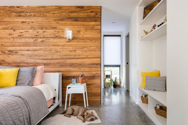 In this modern master bedroom, a wood accent wall adds warmth to the mostly white room, while open shelving and a bench have been built into the wall.