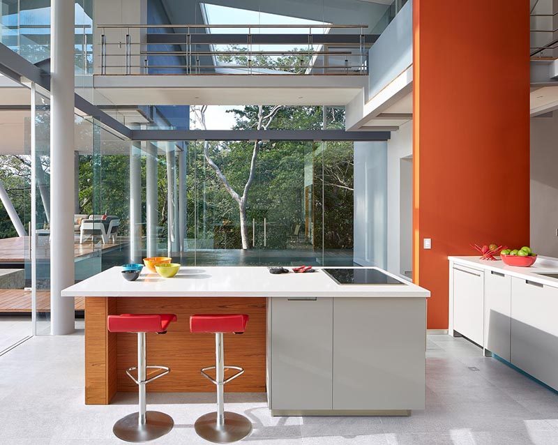 This small and modern kitchen has minimal cabinets, with the island home to the oven and cooktop. A bright orange wall adds a pop of color to the space.