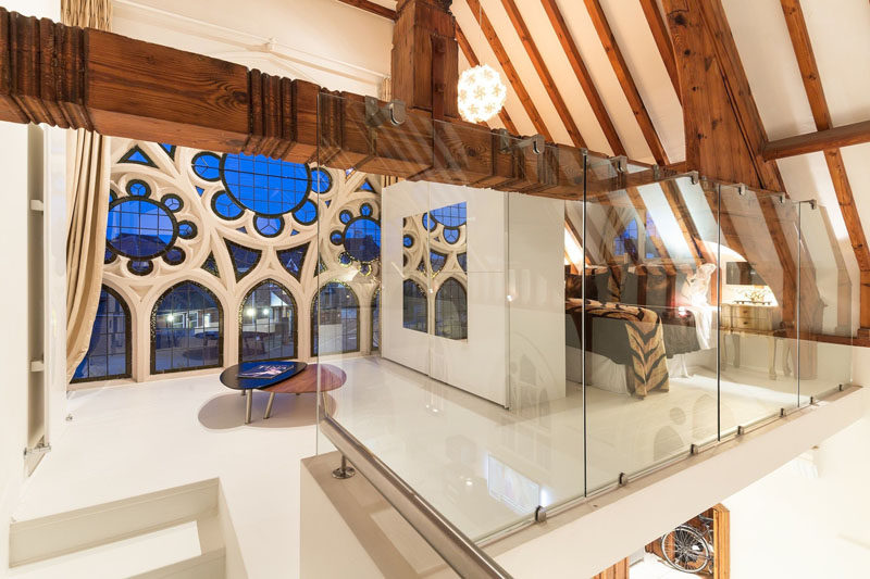 Upstairs and at one end of this renovated church is a bedroom that overlooks the living area below and takes advantage of the light from the original windows. #RenovatedChurch #ChurchConversion #WoodBeams #ModernBedroom #InteriorDesign #Windows