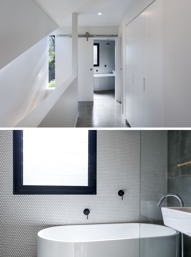 At the top of the stairs in this modern house is a bathroom that's hidden behind a white sliding barn door. The bathroom features a freestanding bathtub with rain shower, an accent wall of penny tiles and matte black accents that match the black window frame. #ModernBathroom #PennyTiles #BathroomIdeas #InteriorDesign #SlidingBarnDoor