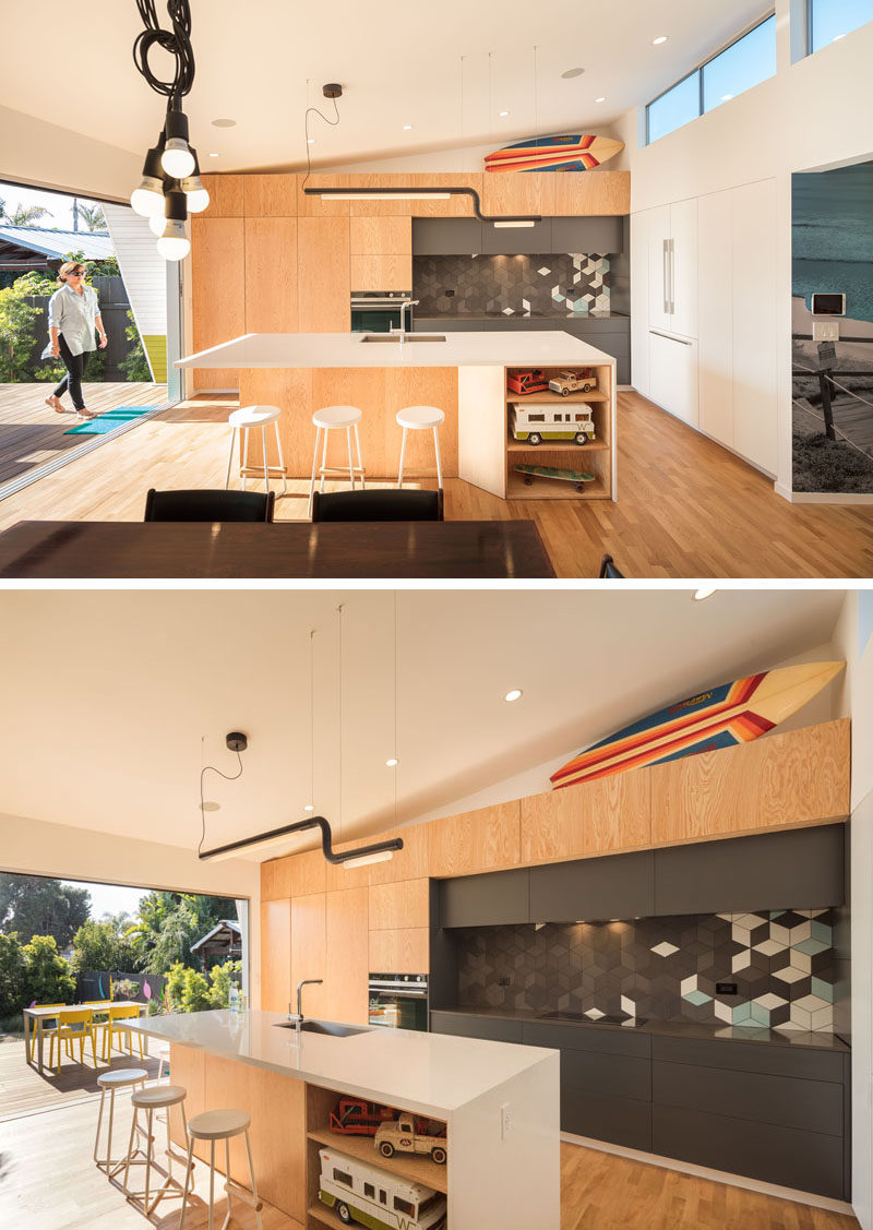 In this modern kitchen, a large island provides ample counter space, while white and wood cabinets provide storage. A section of grey cabinets has a geometric grey, white and blue tile backsplash.