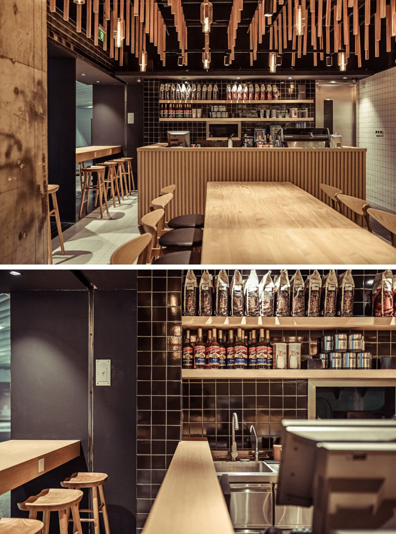 At one end of this modern coffee shop is the service bar which is surrounded by wood, tying in with the other wood elements in the space. Behind the service bar are some open, floating wood shelves on a backdrop of black square tiles.