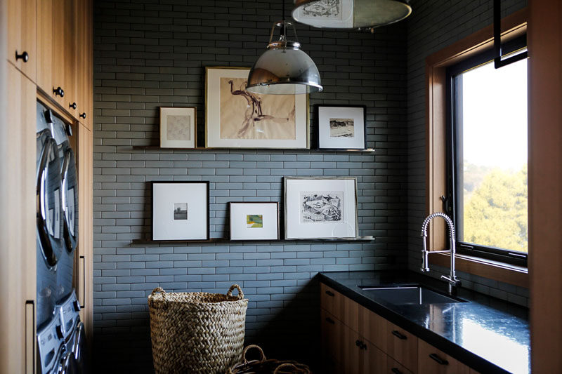 In this modern laundry room, wood cabinets surround the washer and dryer, while grey tile becomes a backdrop for some shelving that displays art.  #LaundryRoomIdeas #LaundryRoom #GreyBrick