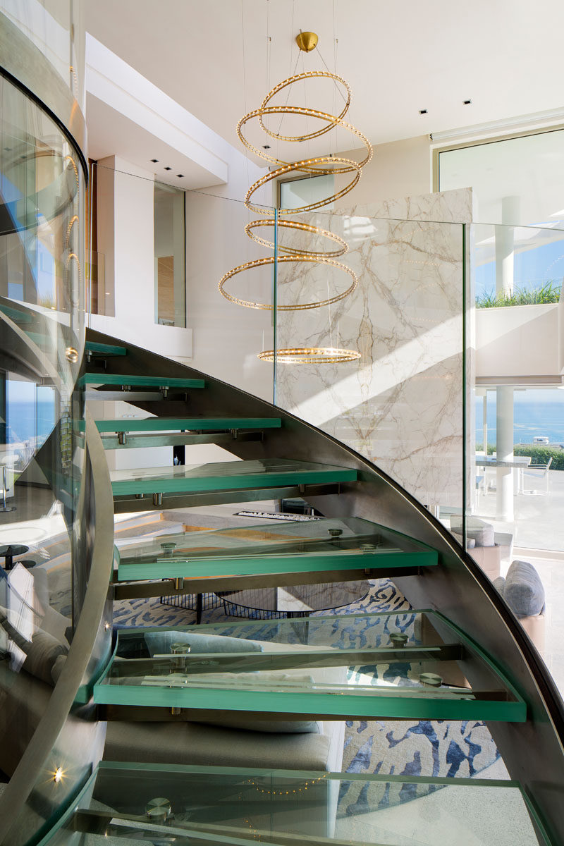 To reach the upper floors of this modern house, there's a triple volume glass lift that's surrounded by a circular glass and steel staircase. An LED metal pendant light fitting by Quasar Citadel compliments the staircase perfectly with its circular cascade design. #SpiralStaircase #GlassStaircase #SculpturalLighting