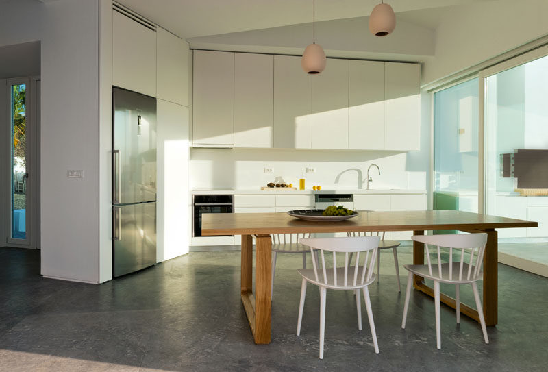 This modern and minimalist kitchen has white cabinets that blend into the walls, while a wood table with white chairs provides a place to eat. #WhiteKitchen #WoodDiningTable #ModernInteriorDesign