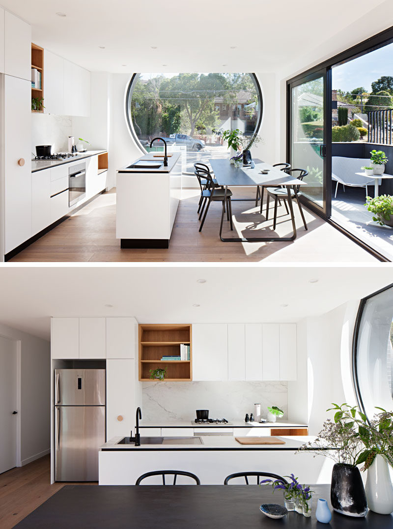 This kitchen is minimal in design, with a long island separating the dining room and a wall of kitchen cabinets. Open wood shelving breaks up the all white cabinets, and a large porthole window floods the room with natural light. #Kitchen #ModernKitchen #WhiteCabinets #DiningRoom #InteriorDesign