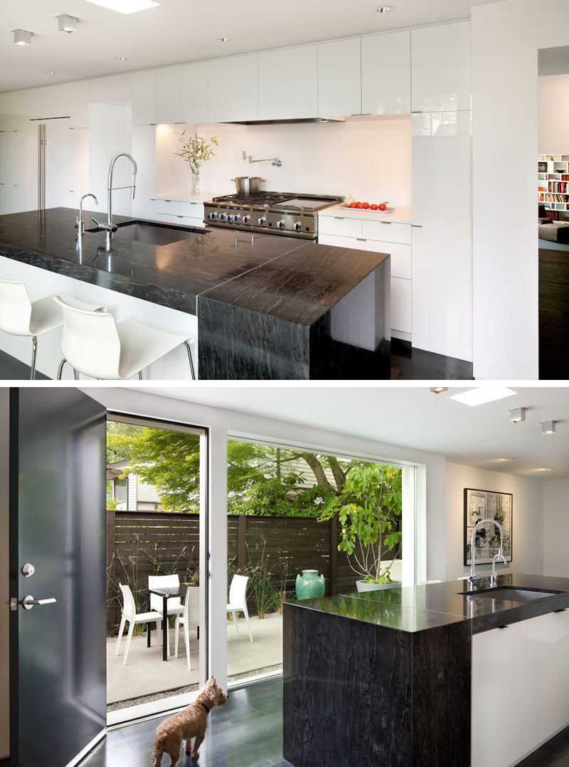 White glossy cabinets and a large island with an additional floating section and dark granite make this kitchen modern in appearance. Off the kitchen is a small patio area for casual outdoor dining. #ModernKitchen #WhiteKitchenCabinets #DarkGranite #InteriorDesign #Patio